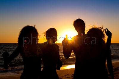 People (two couples) on the beach having a party, drinking and having a lot of fun in the sunset (only silhouette of people to be seen, people having bottles in their hands with the sun shining through)