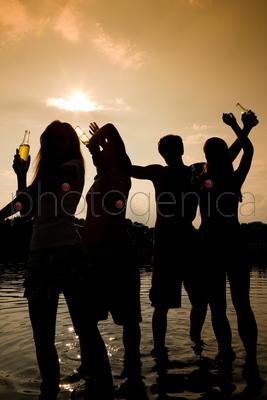 People (two couples) on the beach dancing to music, drinking and having a lot of fun in the sunset - only silhouette of people to be seen